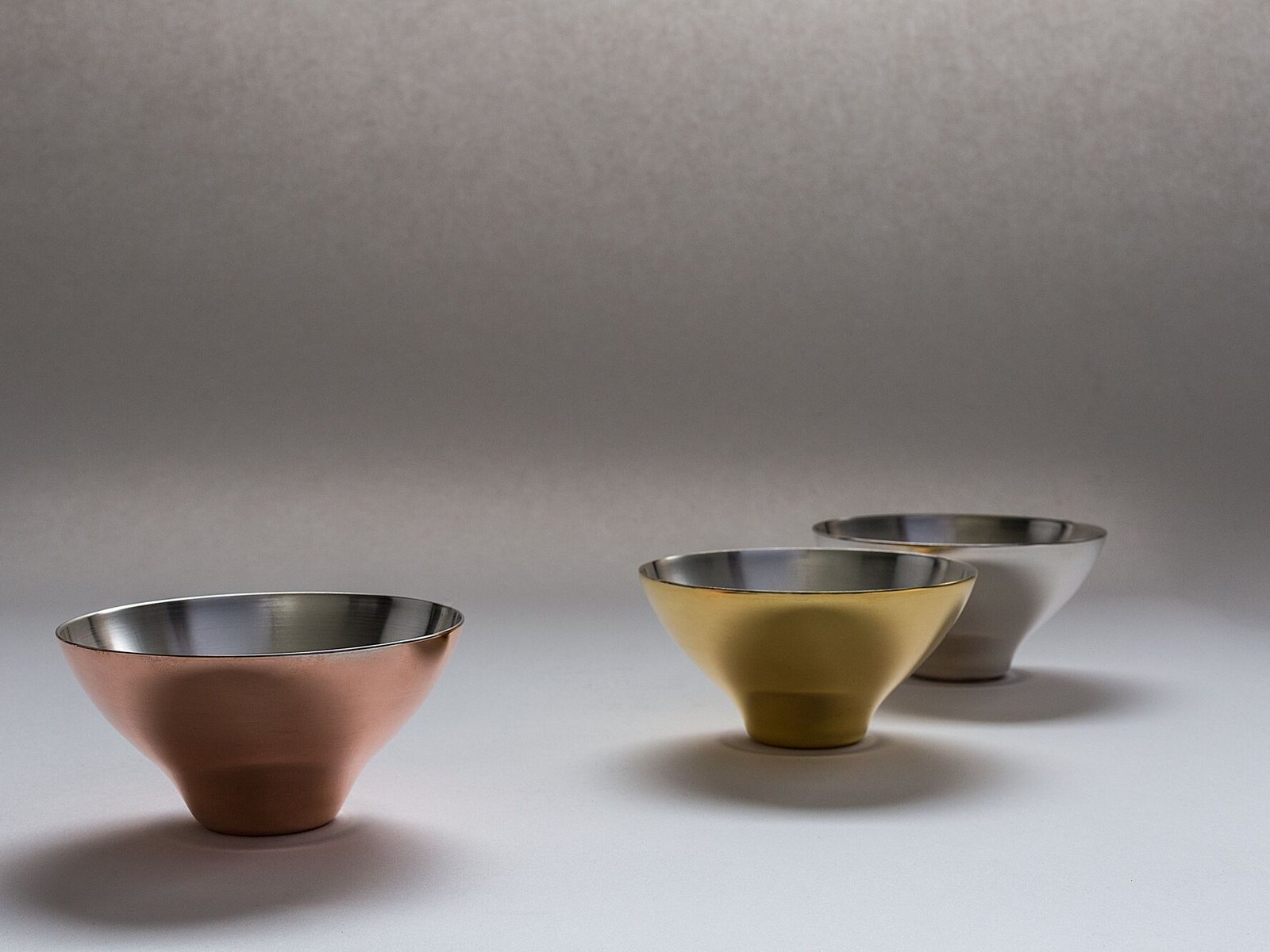 Three spun bowls side by side — copper, brass and steel — on a pale ground