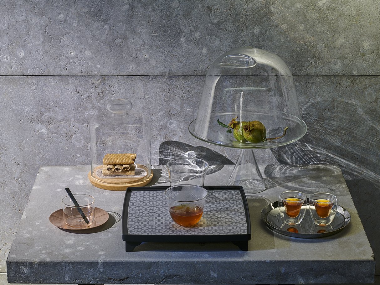Nu glass on a stone table — a dome over fruit, a tumbler of tea on a tray, two double-walled cups on a steel plate