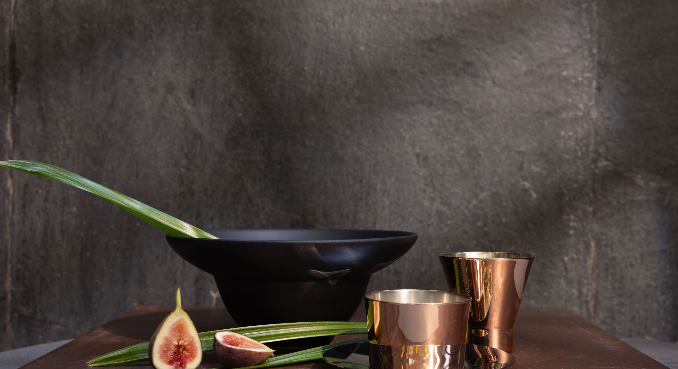 A blackened bowl and two copper cups on a dark ledge, figs and palm alongside