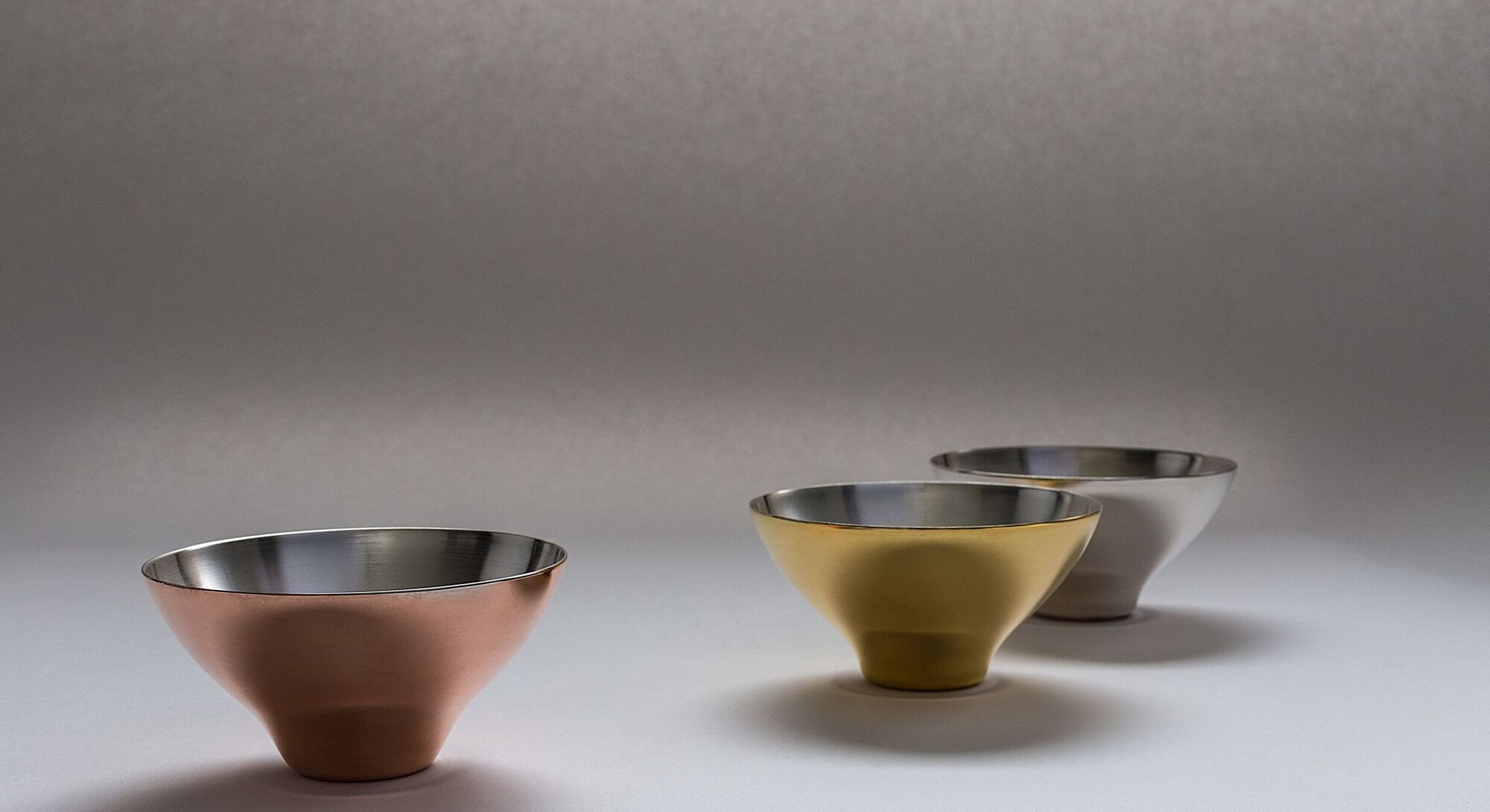 Three spun bowls in copper, brass and steel on a pale ground